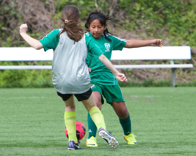 Zwei Mädchen in grünen Jerseys spielen Fußball auf grünem Gras mit einer weißen Bank und Bäumen im Hintergrund.