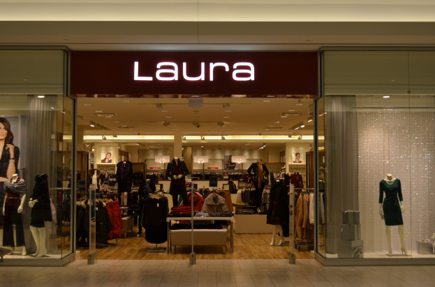 Innenansicht eines Laura-Geschäfts in einem Einkaufszentrum, mit Schaufensterpuppen in modernen Outfits, aufgehängten Kleidungsstücken, einer digitalen Anzeige, einem Geschäftsschild, Deckenbeleuchtung und Fliesenboden.