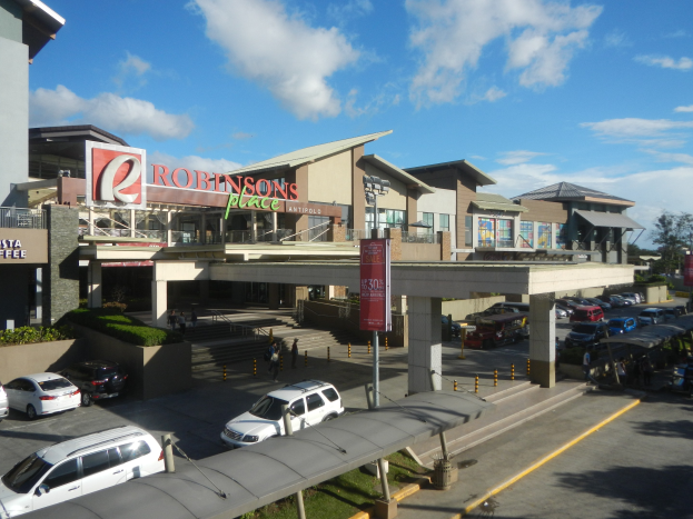 Ein belebtes Einkaufszentrum mit Gebäuden, Geschäftsfronten, Fußgängern, Fahrzeugen, Straßenmöblierung, Grünanlagen und einem bewölkten Himmel.