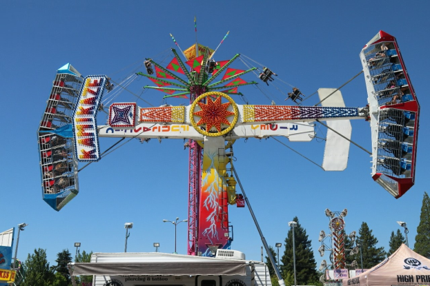 Kirmesfahrt mit sitzenden Menschen auf einer Kirmes, umgeben von Zelten, Bannern, Pfählen, Lichtern und Bäumen unter einem sichtbaren Himmel.