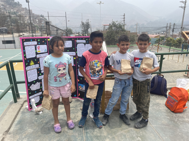 Gruppe von Kindern auf einem Basketballfeld mit Papierbeuteln in den Händen, Beutel und Gegenstände rechts auf dem Boden und eine Tafel mit Text links, vor einem Hintergrund aus Zäunen, Pfosten, Straßenlaternen, Strommasten, Bäumen, Gebäuden, Hügeln und Himmel.