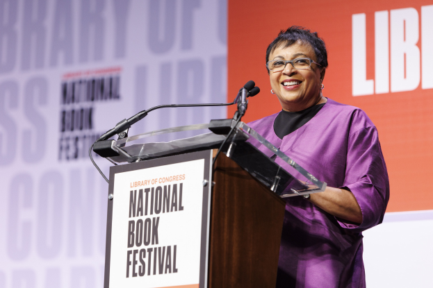 Eine Frau in einem purpurfarbenen Kleid steht an einem Podium mit zwei Mikrofonen und einer angebrachten Tafel vor einem Bildschirm, mit einem orange-weißen "National Book Festival"-Banner im Hintergrund.