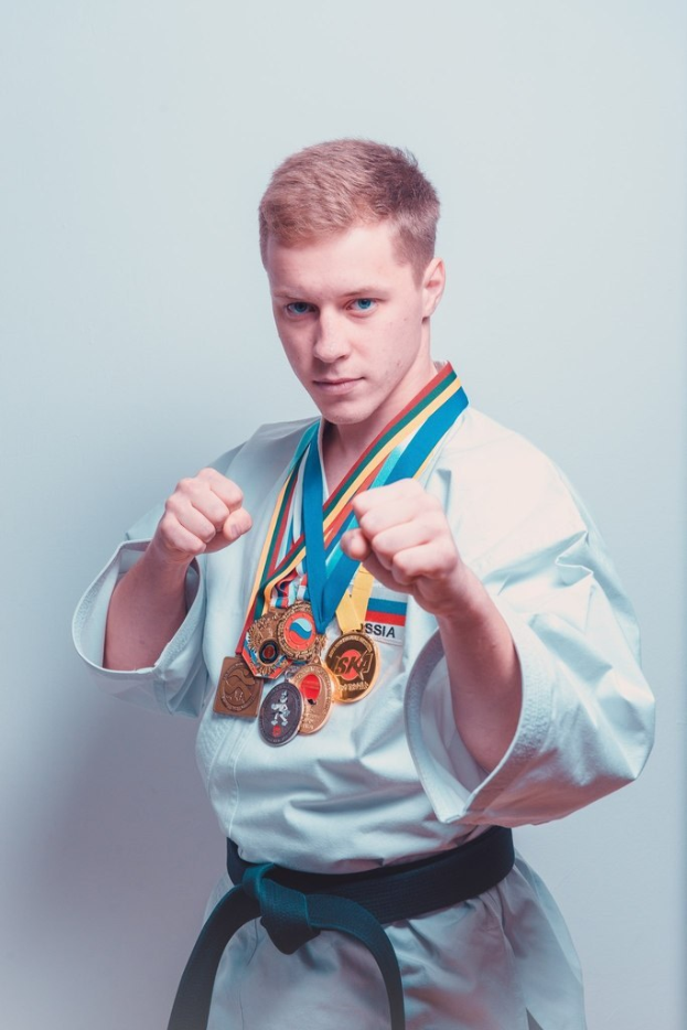 Ein junger Mann in einem Karate-Anzug mit Medaillen um den Hals, der einen schwarzen Gürtel trägt und vor einer Wand steht.