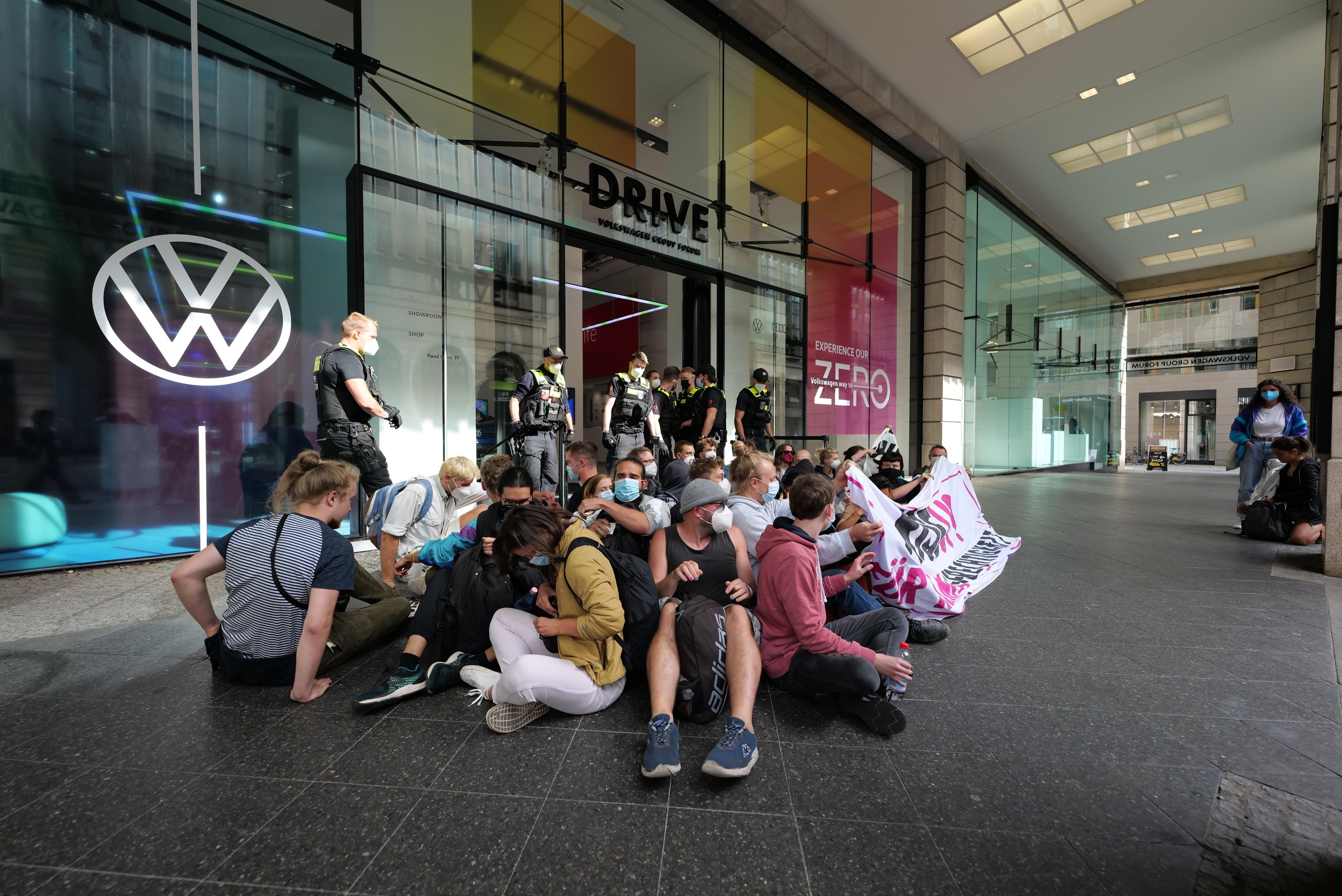 Eine Gruppe von Menschen protestiert vor einer Volkswagen-Niederlassung, einige halten ein Transparent, mit dem Glasgebäude und Logo im Hintergrund.