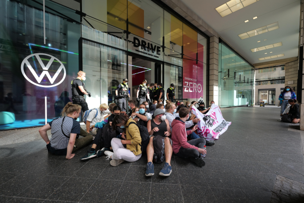 Eine Gruppe von Menschen protestiert vor einer Volkswagen-Niederlassung, einige halten ein Transparent, mit dem Glasgebäude und Logo im Hintergrund.