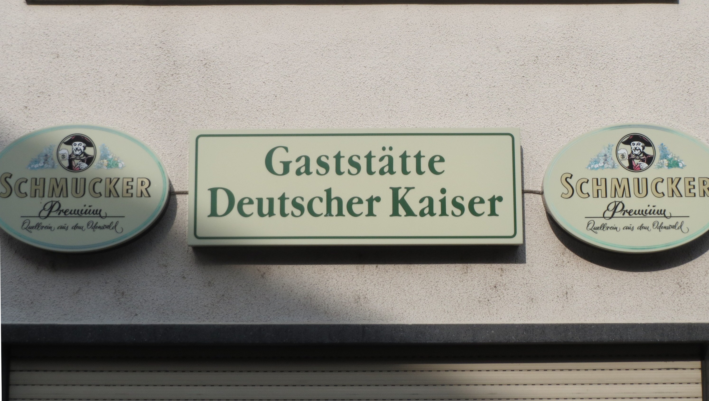 Building with three wall-mounted boards displaying the text "Gaststätte Deutscher Kaiser Schmucker" and a shutter at the bottom.
