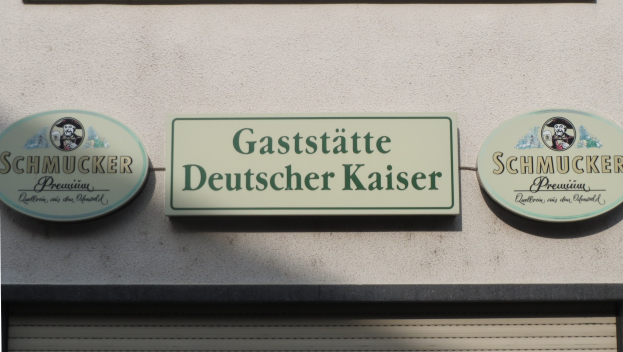 Building with three wall-mounted boards displaying the text "Gaststätte Deutscher Kaiser Schmucker" and a shutter at the bottom.