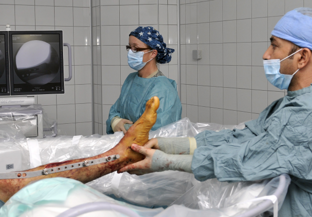 Zwei Chirurgen in Kitteln und Masken führen einen Eingriff am Bein eines Patienten in einem Operationssaal durch, mit medizinischen Geräten und einer Wand im Hintergrund.