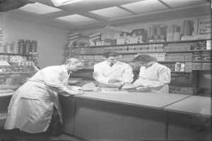 Drei Männer in weißen Kitteln stehen an einem Tresen in einer Apotheke, mit Papieren und Gegenständen darauf und Regalen im Hintergrund, dargestellt in Schwarz-Weiß.