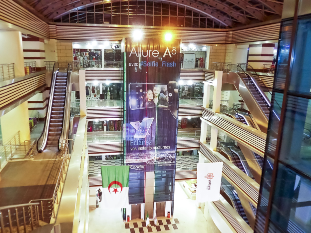 Ein belebtes Einkaufszentrum mit Rolltreppen, Geländern, Bannern, Lichtern und einer Decke, mit einigen Menschen.