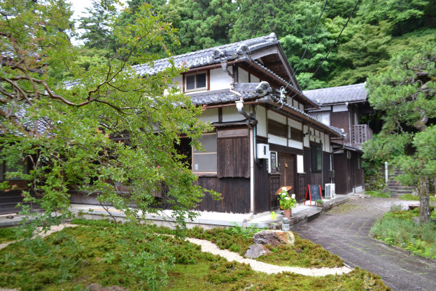 Traditionelles japanisches Haus umgeben von dichtem Waldgrün mit einer Straße, die darauf führt, unter einem klaren Himmel.