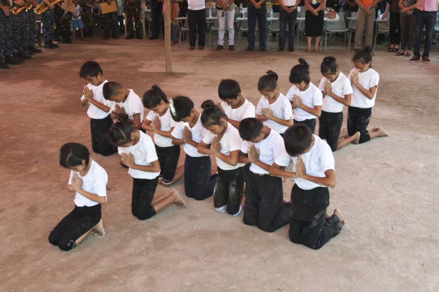 Gruppe von Kindern kniend vor einer Menge mit Musikinstrumenten, mit Stühlen und einem Pfahl in einer Schulumgebung.
