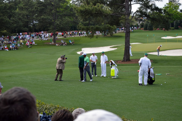 Eine Gruppe von Menschen, die Golf auf einem Kurs mit Ausrüstung und Zuschauern spielt, mit Bäumen im Hintergrund und einem Fotografen, der die Szene aufnimmt.