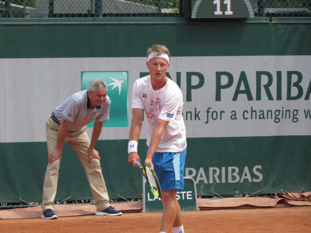 Zwei Männer spielen Tennis auf einem Sandplatz, wobei einer ein weißes T-Shirt und blaue Shorts trägt und einen Schläger hält, ein grünes Banner mit Text dahinter und ein Zaun und eine Nummernanzeige im Hintergrund.