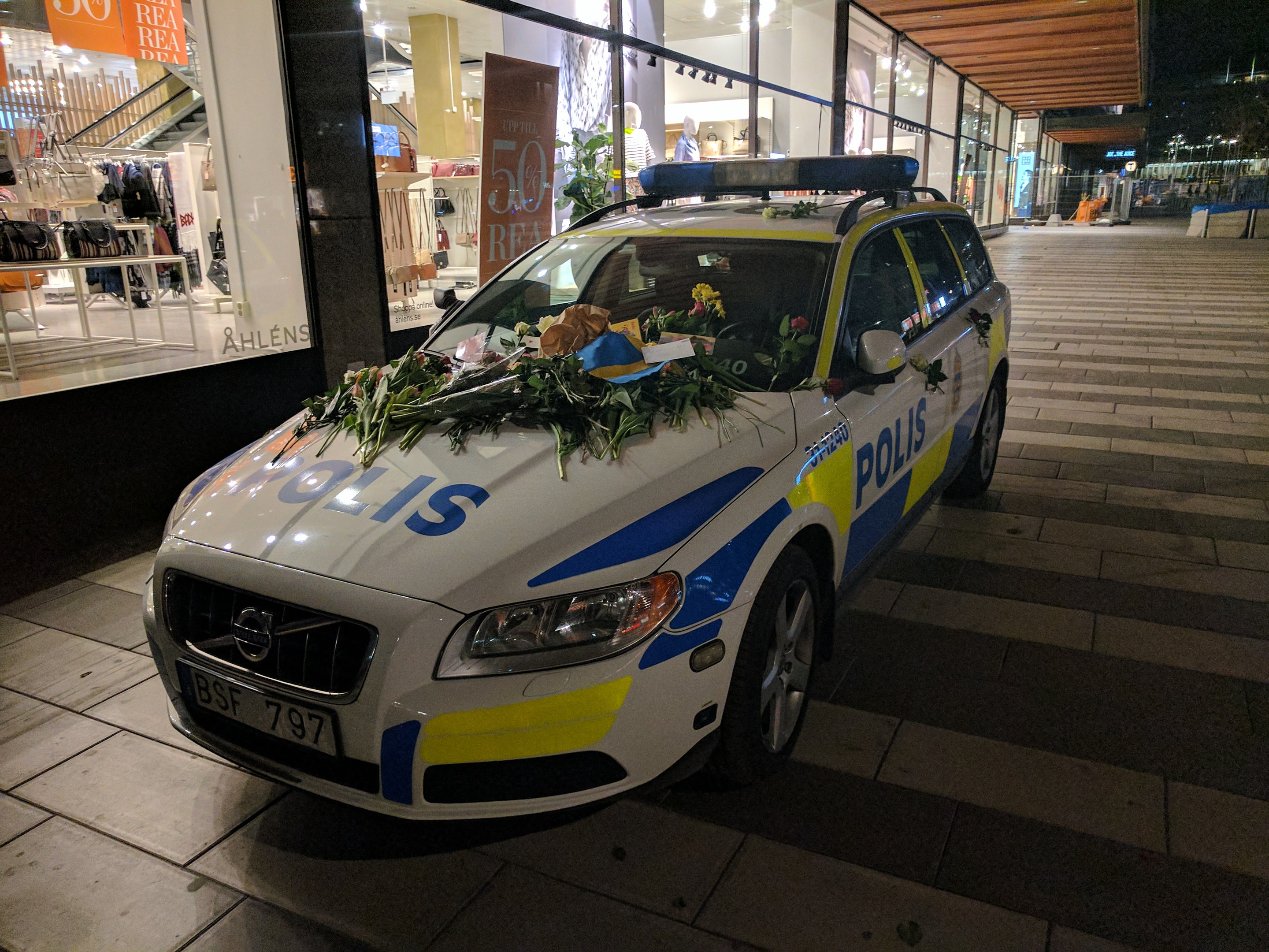Ein mit Blumen und Blättern geschmückter Polizeiwagen parkt nachts vor einem Geschäft mit Glaswänden, das Puppen in Kleidern und beleuchtete Schilder zeigt.