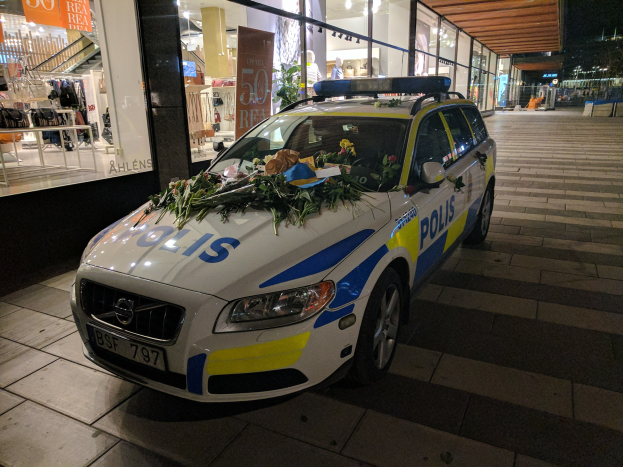 Ein mit Blumen und Blättern geschmückter Polizeiwagen parkt nachts vor einem Geschäft mit Glaswänden, das Puppen in Kleidern und beleuchtete Schilder zeigt.
