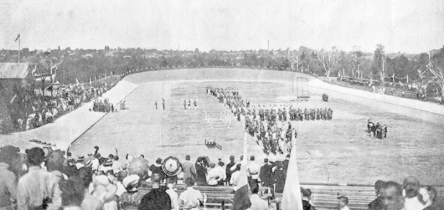Schwarzes und weißes Foto von Zuschauern bei einem Pferderennen in einem Stadion, einige sitzen auf Bänken und andere stehen, mit Reitern und Pferden auf der Bahn, Bäumen im Hintergrund und einem klaren Himmel.