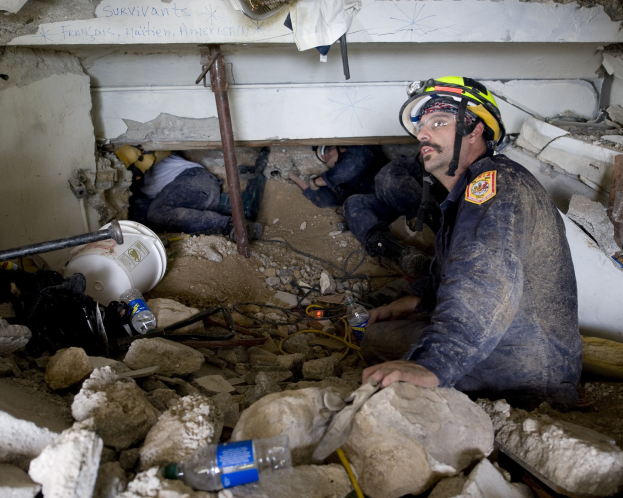 Feuerwehrmann mit Helm, der in Trümmern eines eingestürzten Geb├Ąudes sitzt, umgeben von Steinen, Flaschen und anderen Gegenst├Ąnden. Im Hintergrund eine Wand mit etwas darauf geschrieben, wahrscheinlich der Name des Geb├Ąudes, das Ort eines israelischen Luftangriffs ist.