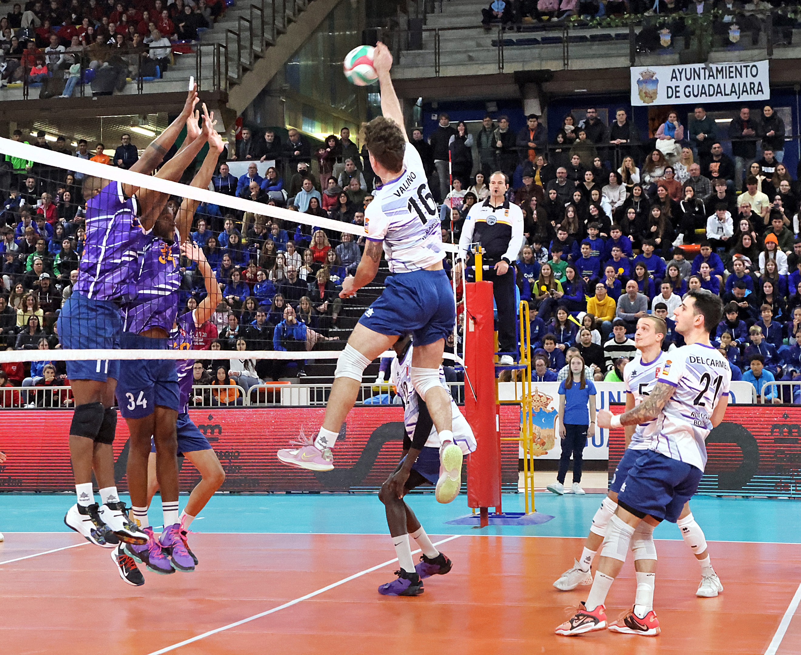 Eine Gruppe von Männern spielt Volleyball auf einem Court mit einem Netz, während Zuschauer auf Stühlen im Hintergrund sitzen, mit Geländern, Stufen und Bannern im Hintergrund.