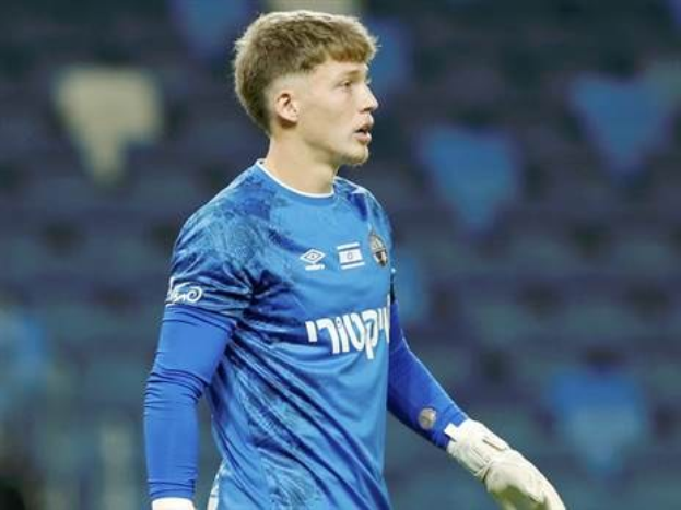 Ein junger Mann in einem blauen Torwarttrikot steht auf einem Fußballfeld während eines Spiels, trägt ein blaues T-Shirt und weiße Handschuhe.