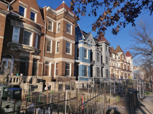 Reihe von Häusern in einer Wohngegend mit einem Zaun, Fenstern, Bäumen, Mülltonnen und sichtbarem Himmel im Hintergrund.