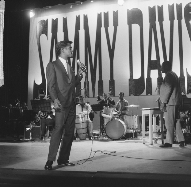 Schwarz-weißes Foto von Sammy Davis Jr., der auf der Bühne des Apollo Theater auftritt und ein Mikrofon hält, während er einen Blazer und eine Krawatte trägt und von einer Band und Scheinwerfern unterstützt wird.