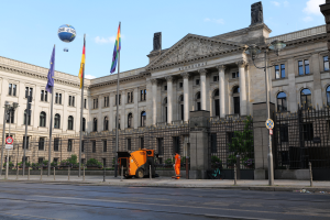 Das Reichstagsgebäude in Berlin, Deutschland, ein großes Gebäude mit zahlreichen Fenstern, umgeben von Straßenelementen, Fahrzeugen, Menschen und Bäumen unter einem bewölkten Himmel.