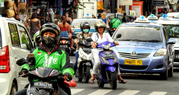 Eine Gruppe von Motorradfahrern fährt auf einer vielbefahrenen Straße neben Autos und anderen Fahrzeugen, wobei einige Fahrer Helme tragen und Gebäude, Bäume und Geschäfte im Hintergrund zu sehen sind.