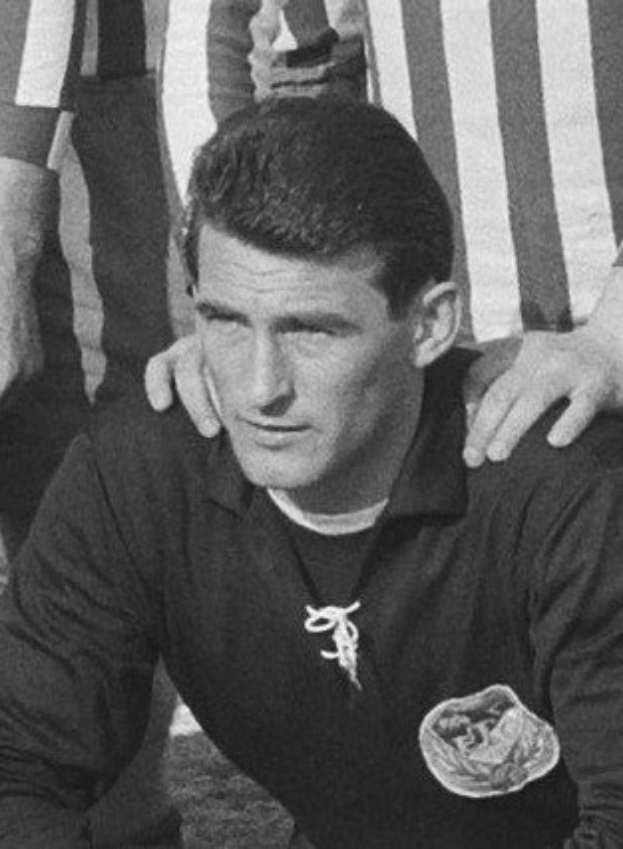 Black and white photo of a man sitting on a grassy soccer field wearing a t-shirt and a locket, with two people standing behind him.