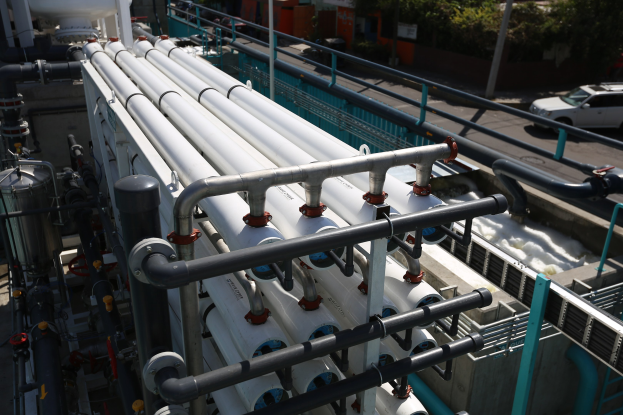 Large group of pipes and valves on a building roof at a wastewater treatment plant, with railings, vehicles, trees, and surrounding buildings in the background.