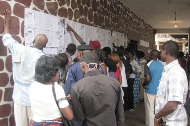 Eine Gruppe von Menschen steht vor einer Wand mit Papieren, beleuchtet von Deckenlampen, einige tragen Taschen, was auf einen Abstimmungsprozess hinweist.