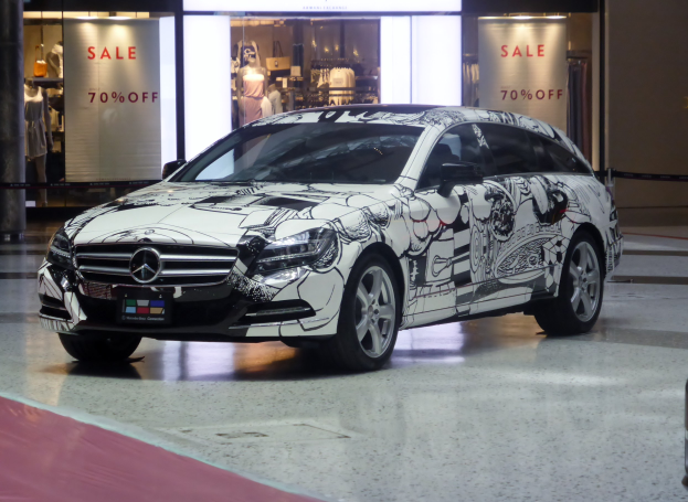 Ein silberner Mercedes-Benz CLS Shooting Brake vor einem Geschäft mit Schaufensterpuppen in Kleidern im Hintergrund, einer Säule auf der linken Seite und einer Wand auf der rechten Seite.