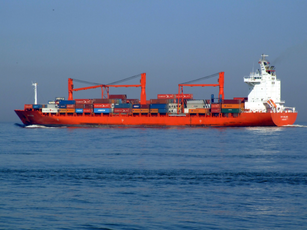 Ein großes rotes und weißes Containerschiff segelt auf dem Meer und trägt zahlreiche Container bei klarem blauem Himmel.
