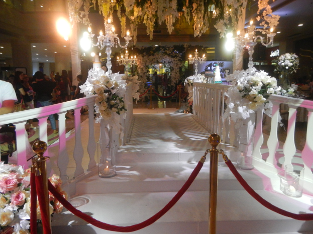 Eine Hochzeitszeremonie in einem Einkaufszentrum mit weißem Teppich, Geländern, dekorativen Pfosten mit Seilen, Blumenarrangements, Kronleuchtern, Beleuchtung und zahlreichen Gästen, einige mit Taschen, unter einer Decke mit Lichtern.