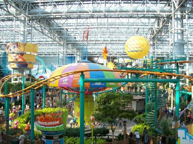 Großes Indoor-Wasserpark mit Achterbahn umgeben von Menschen, Bäumen, Pflanzen und Beleuchtungsstrukturen in einer lebendigen Decke.