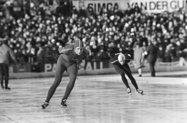 Zwei Eisschnellläufer in Schwarz-Weiß-Aktion auf einer Eisbahn, mit Helmen und Skating-Schuhen, mit Zuschauern und Tafeln im Hintergrund.