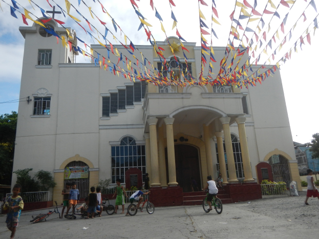 Eine Gruppe von Menschen, die vor einer weißen Kirche mit architektonischen Details wie Fenstern, Säulen, Bögen, Türen und einem Kreuzsymbol Fahrrad fahren, vor einem bewölkten Himmel mit Bäumen, Pflanzen, einem Zaun und einer Fahne im Hintergrund, mit bunten Fahnen, die an einem Seil über ihnen befestigt sind.