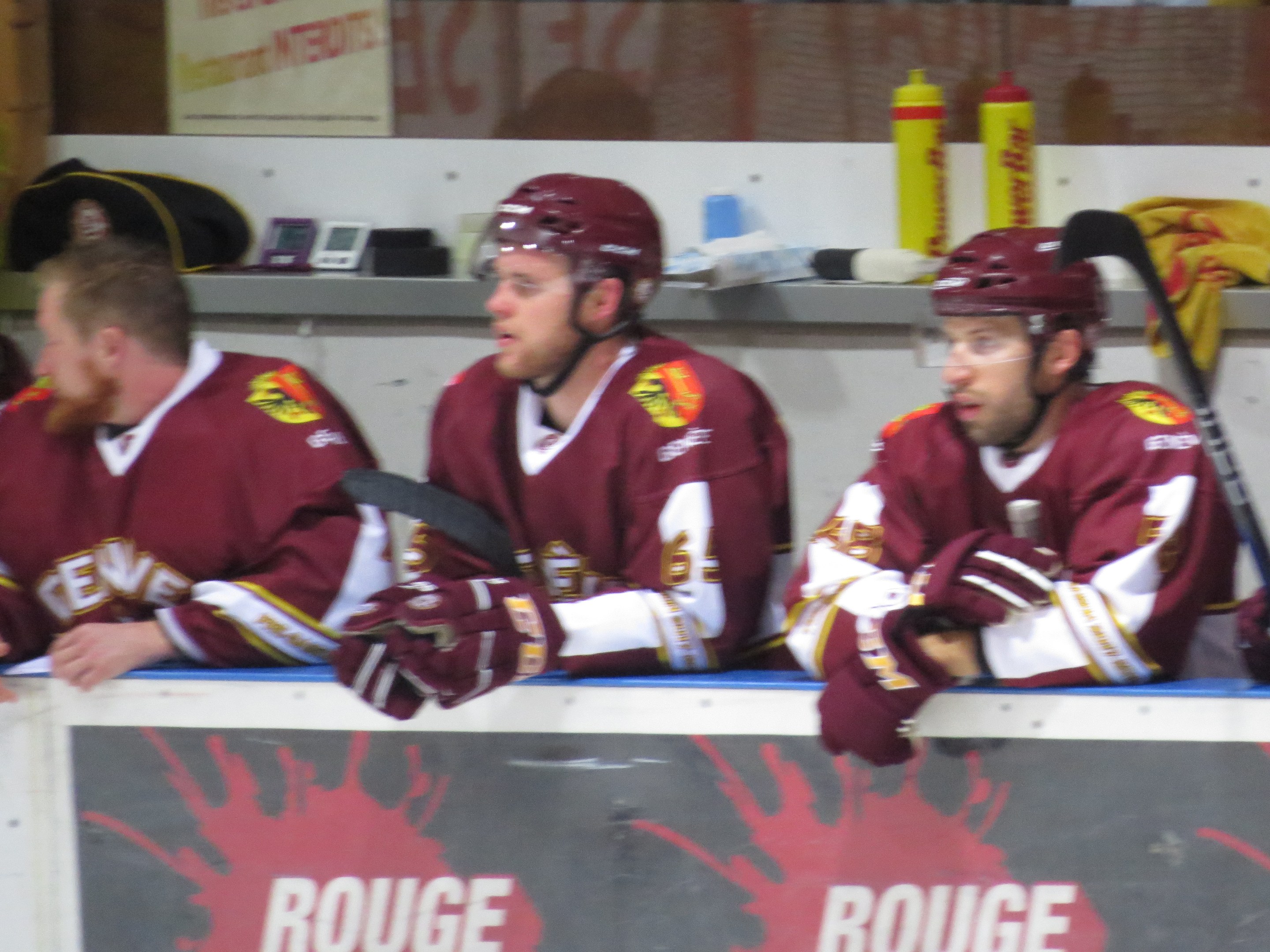 Drei Hockey-Spieler in rotbraunen Jerseys und Helmen sitzen auf einer Bank und halten Hockey-Schläger, vor ihnen ein Schild und weitere Personen im Hintergrund.