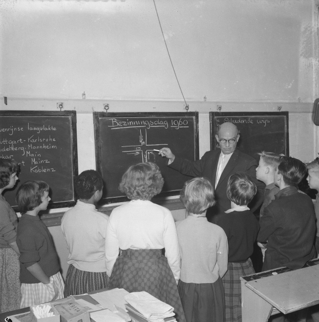 Schwarzes und weißes Foto eines Lehrers, der vor einer Tafel mit einem Marker steht und sitzende Kinder an einem Tisch mit Büchern und Gegenständen unterrichtet, mit einem an der Wand montierten Lautsprecher auf der rechten Seite.