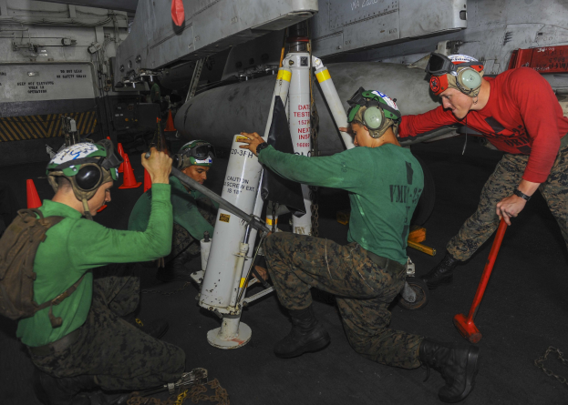 Eine Gruppe von Männern in grünen Hemden und Helmen arbeitet an einer Rakete, umgeben von Verkehrskegeln und anderen Gegenständen auf dem Boden, mit einem Flugzeug im Hintergrund.
