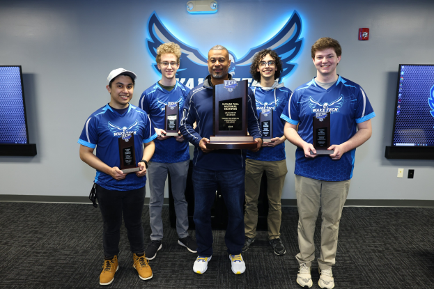 Fünf Männer halten Trophäen in einem Raum mit NCAA-Fall-Championship-Logos und Fernsehgeräten im Hintergrund.