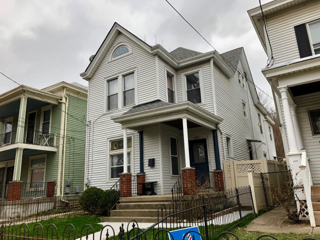 Reihe von Häusern mit Fenstern, Türen, Säulen, Geländern, Stufen und Dächern, umgeben von einem Metallzaun, Gras, Pflanzen, Bäumen und einem bewölkten Himmel, mit einem Mietschild am Boden, das die Adresse und die Immobilien Details auflistet.