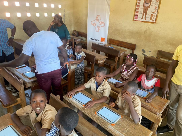 Children sitting at desks using tablets in a classroom with adults standing nearby, a banner, and a poster on the wall.