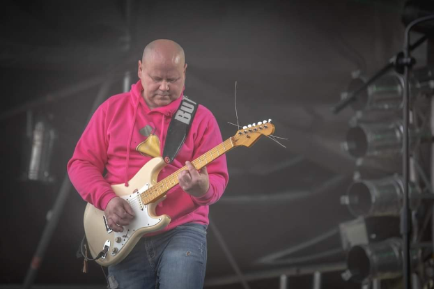 Ein Mann in einem pinken Sweatshirt spielt eine Gitarre auf der Bühne eines Musikfestivals, umgeben von Metallstangen und anderen Gegenständen.
