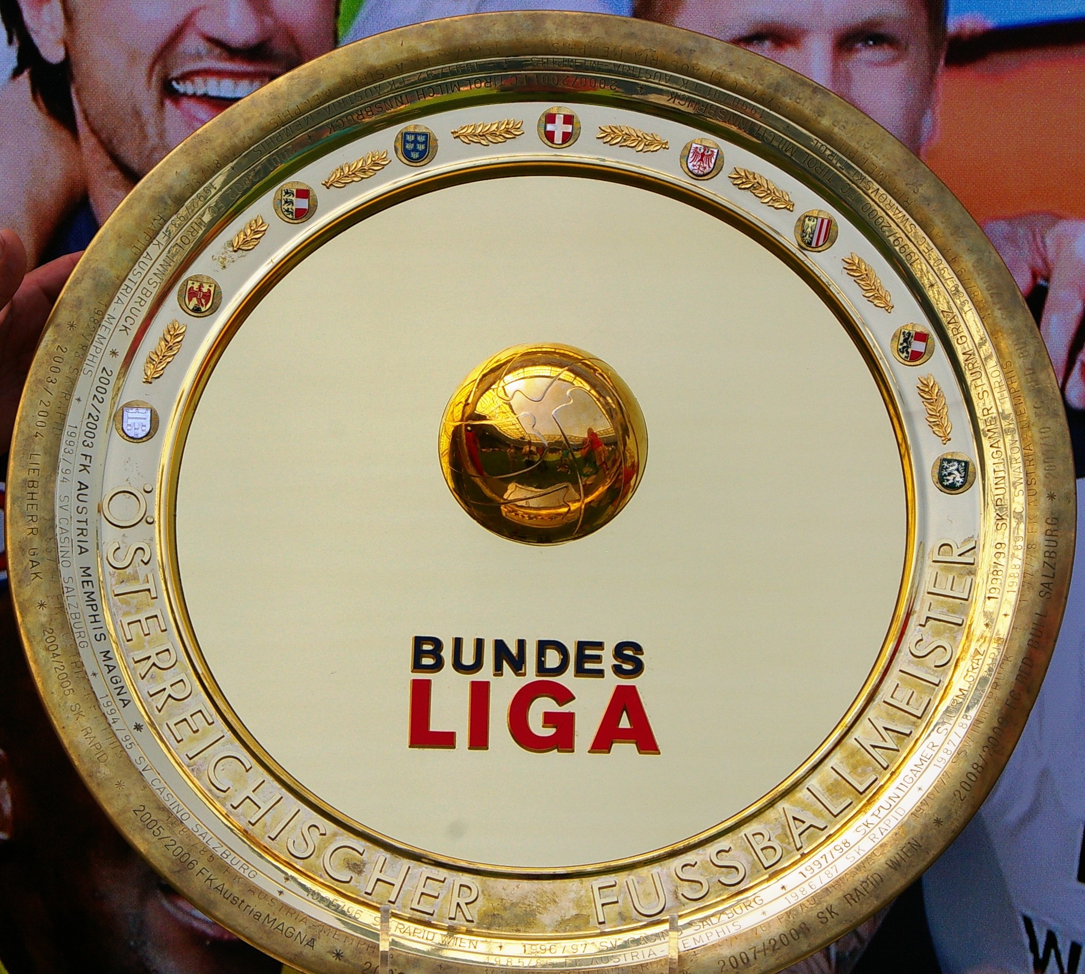 Goldener Schild mit 'Bundesliga'-Text, gehalten von einem Mann, mit einem Banner im Hintergrund, das drei Personen zeigt.