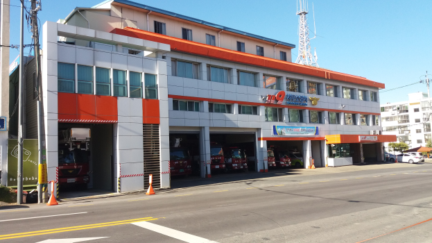 Feuerwache mit Feuerwehrauto davor, umgeben von Gebäuden, Fahrzeugen, Verkehrskegeln, Strommasten, Kabeln, Schildern, Bäumen und einem klaren Himmel.