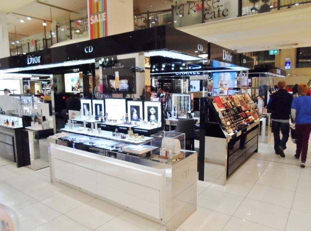 Ein hell erleuchteter Kosmetikladen mit Regalen und Verkaufstischen, auf denen verschiedene Make-up- und Pflegemittel ausgestellt sind, mit mehreren Einkäufern, die die Gänge durchstreifen.