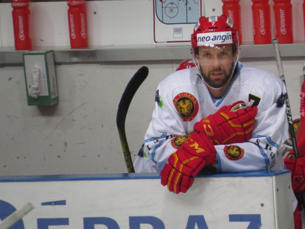 Hockeyspieler sitzt auf der Bank und trägt einen Helm, Handschuhe und hält einen Hockeystock, mit Flaschen und Gegenständen im Hintergrund.