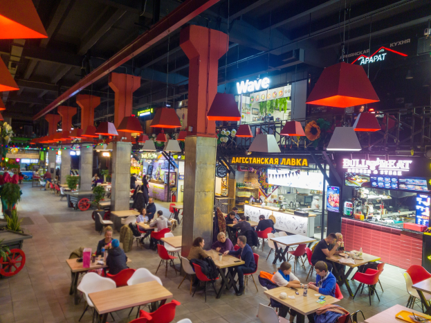 Innenansicht eines belebten Restaurants mit Gästen an Tischen, Flaschen und Gläsern auf den Oberflächen, Deckenleuchten und Säulen, einem Pflanzenwagen auf der linken Seite und Geschäftsfronten im Hintergrund.