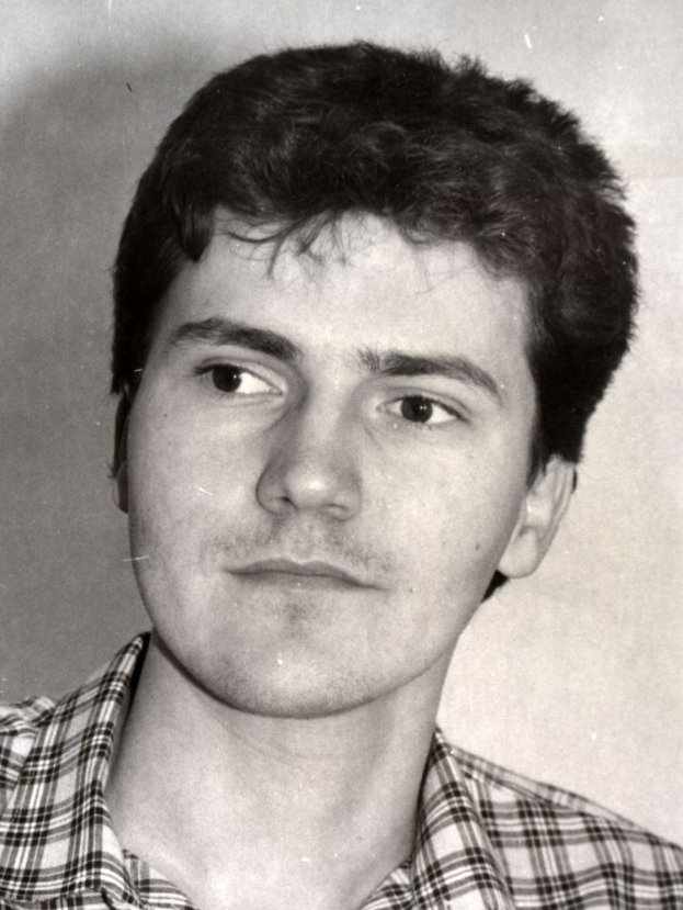 Schwarzes und weißes Foto eines Mannes in einem karierten Hemd mit kurzen dunklen Haaren, der direkt in die Kamera schaut und leicht geöffnete Lippen hat.